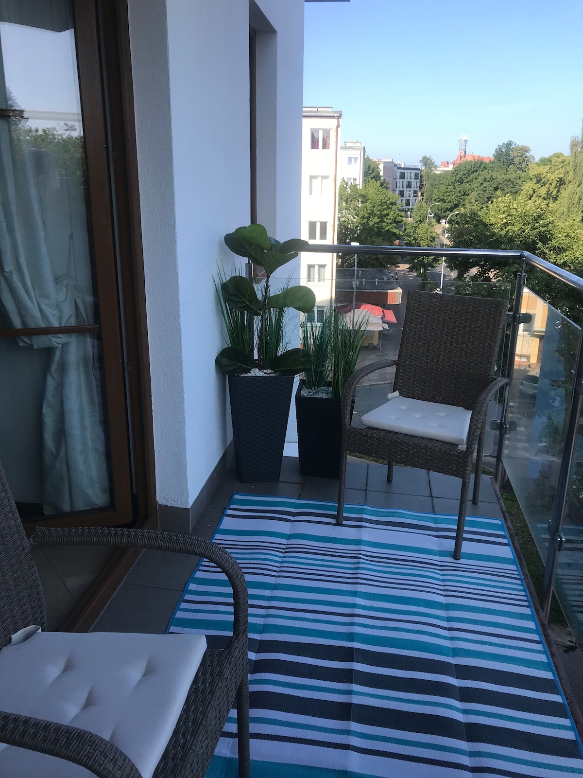 A balcony features two woven chairs beside a small, rectangular outdoor rug. Potted plants provide greenery, enhancing the open space. The view includes street scenes and trees, with clear blue skies above, creating a pleasant setting for relaxation.