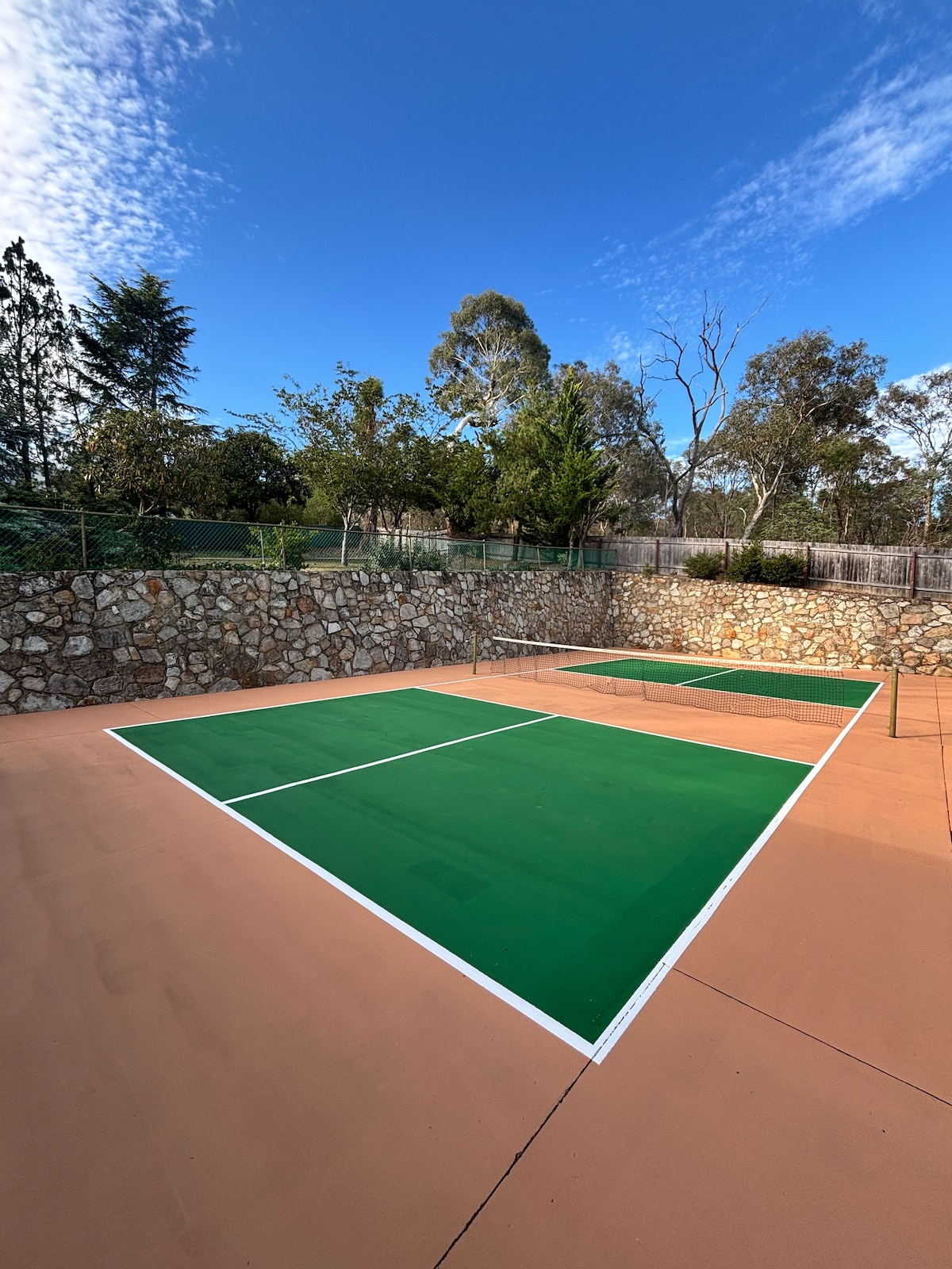 A well-maintained half-sized tennis court is visible, featuring a bright green playing surface bordered with white lines. The surrounding area is lined with a stone wall and trees, contributing to the natural setting under a clear blue sky.