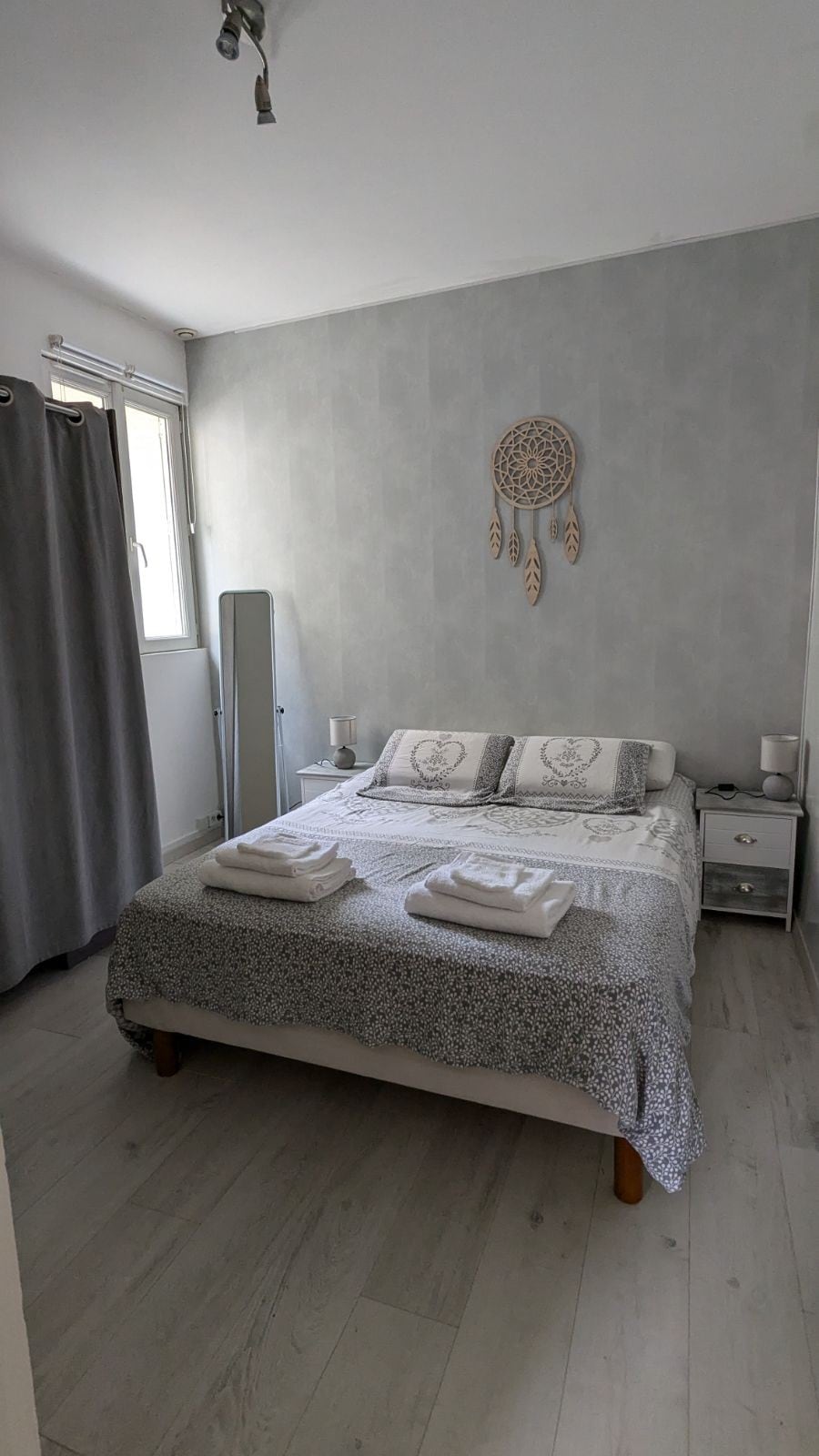 A tranquil bedroom is presented with a spacious bed adorned with decorative pillows and fresh linens. Natural light enters through a window beside gray curtains, while a mirror and bedside table complement the minimalistic decor.