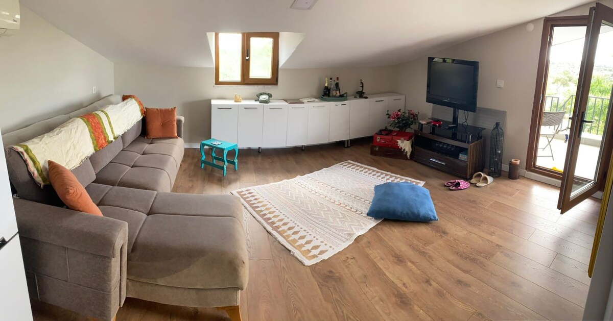 An airy living room features a spacious gray sofa adorned with decorative cushions. A coffee table with a colorful accent sits on a patterned rug. Large windows provide natural light and reveal access to a balcony, enhancing the open feel of the space.