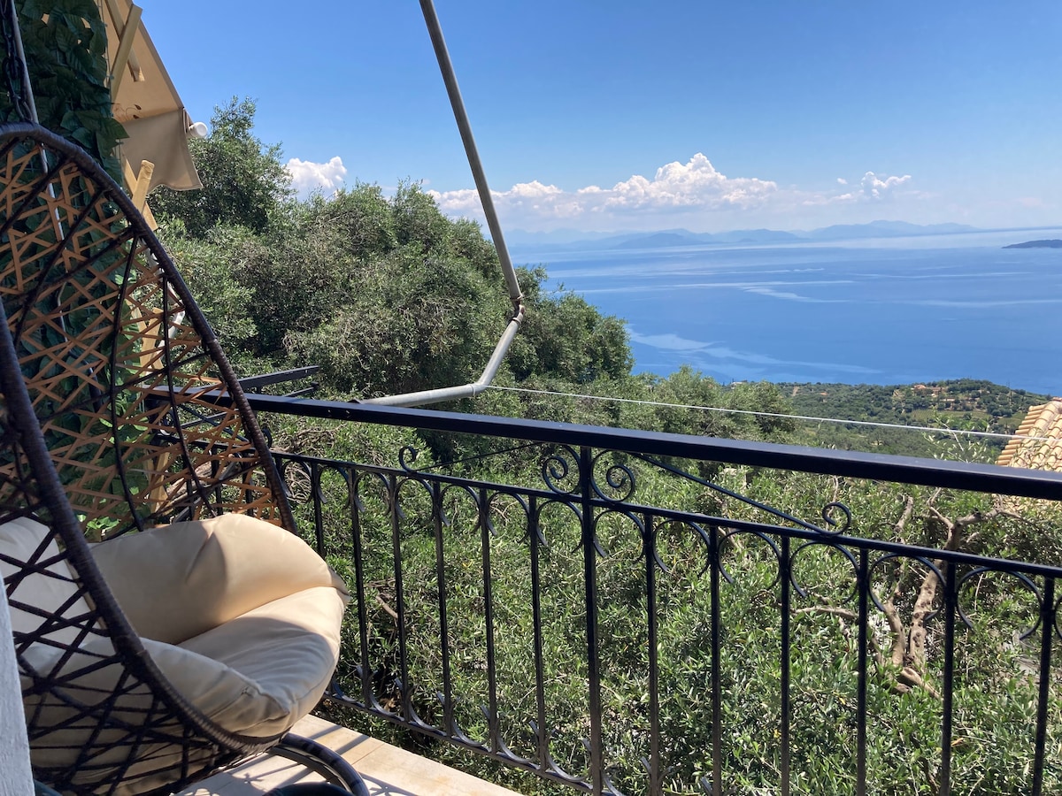 A comfortable chair is positioned on a balcony, surrounded by green foliage. The expansive view of the Ionian Sea is visible, with distant islands and clouds creating a serene backdrop. The railing features intricate designs that complement the natural setting.