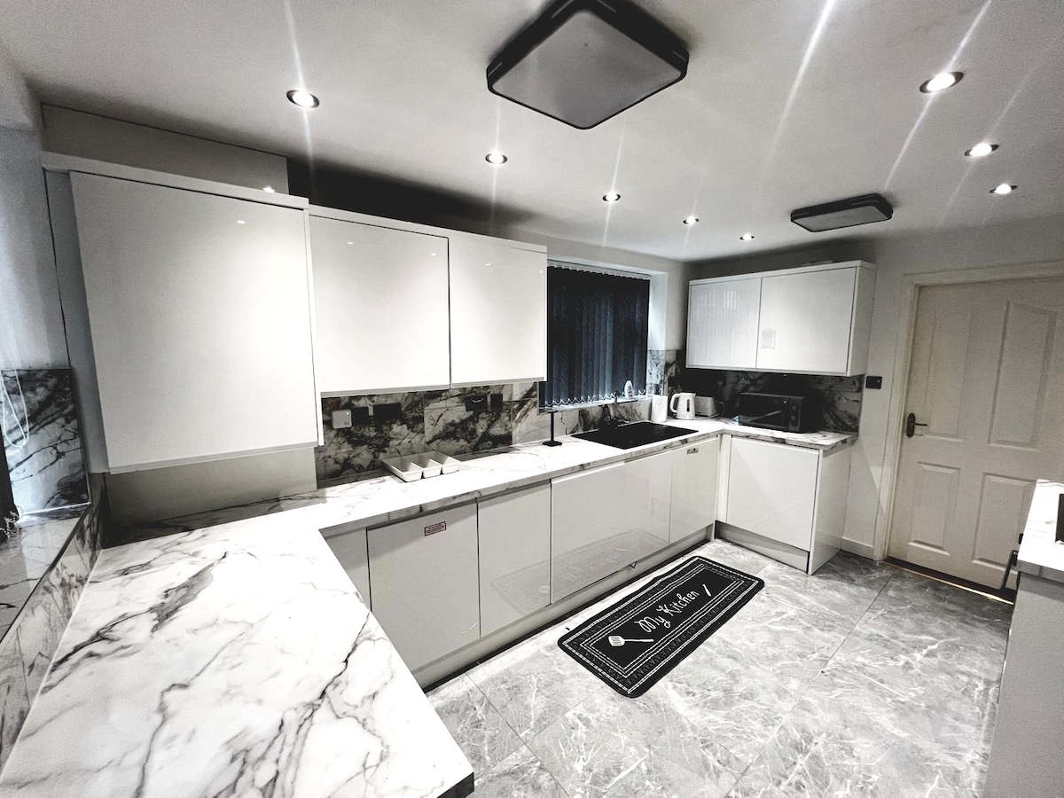 A modern kitchen features sleek white cabinetry complemented by striking marble countertops. Under-cabinet lighting highlights the work surfaces, while stainless steel appliances add functionality. A dark rug provides contrast against the light tile flooring, and a window allows natural light to enhance the space.