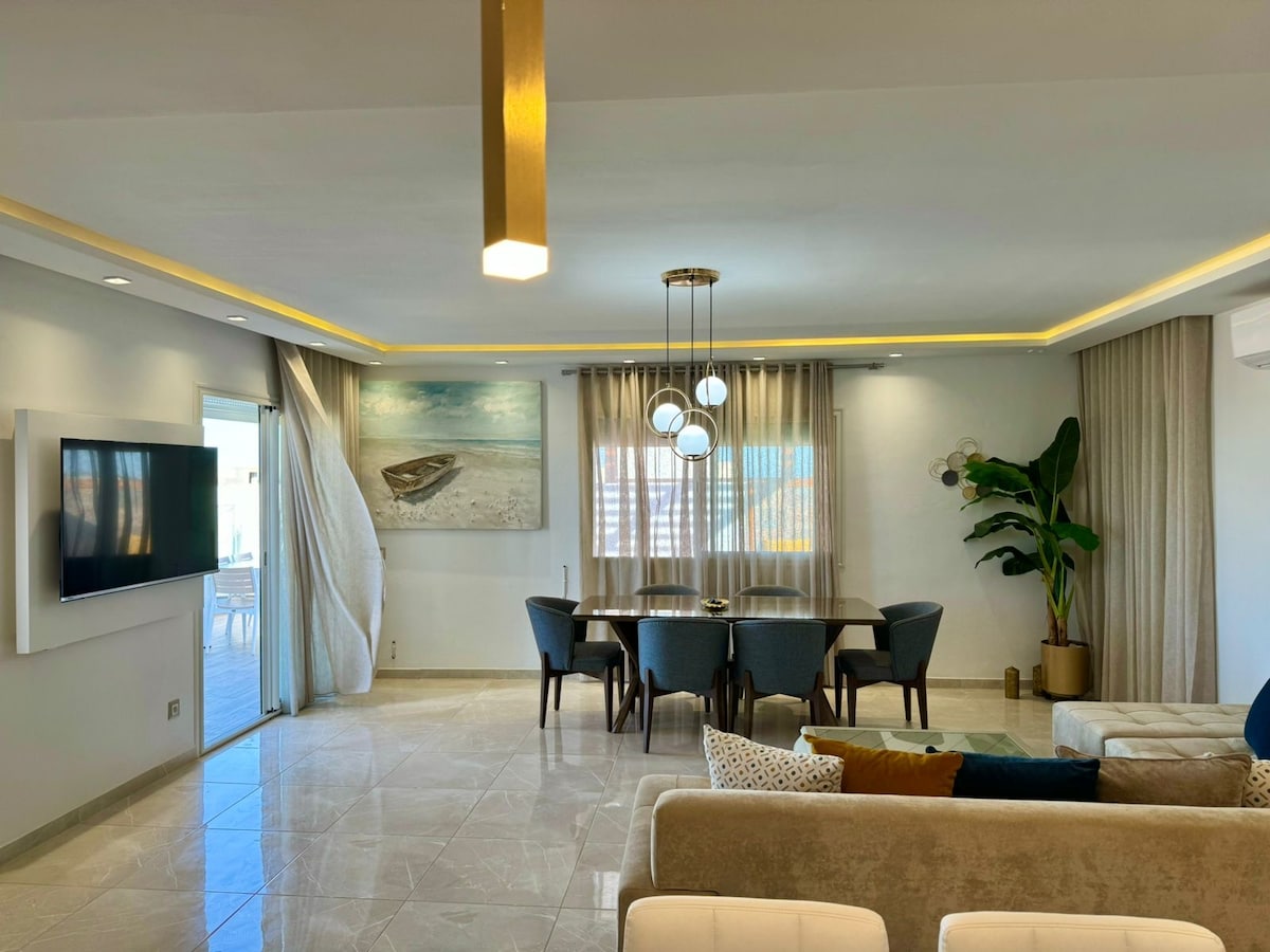A modern living area features a large window allowing natural light to illuminate the space. A dining table with several upholstered chairs is positioned near the window. An artwork of a boat decorates the wall, complemented by a potted plant in the corner.