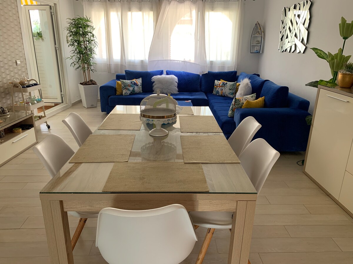 A bright and airy living area features a large blue sectional sofa adorned with decorative pillows. A modern dining table, surrounded by white chairs, sits at the center, complemented by light-colored placemats. Natural light filters through sheer curtains, creating an inviting ambiance.