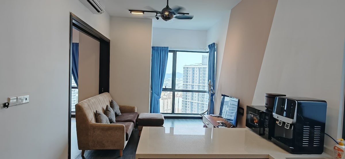 A modern living space is complemented by large windows, providing natural light and city views. A comfortable sofa is positioned in front of a television, with a dining table visible in the foreground. Dark curtains frame the windows, contributing to the overall spaciousness.