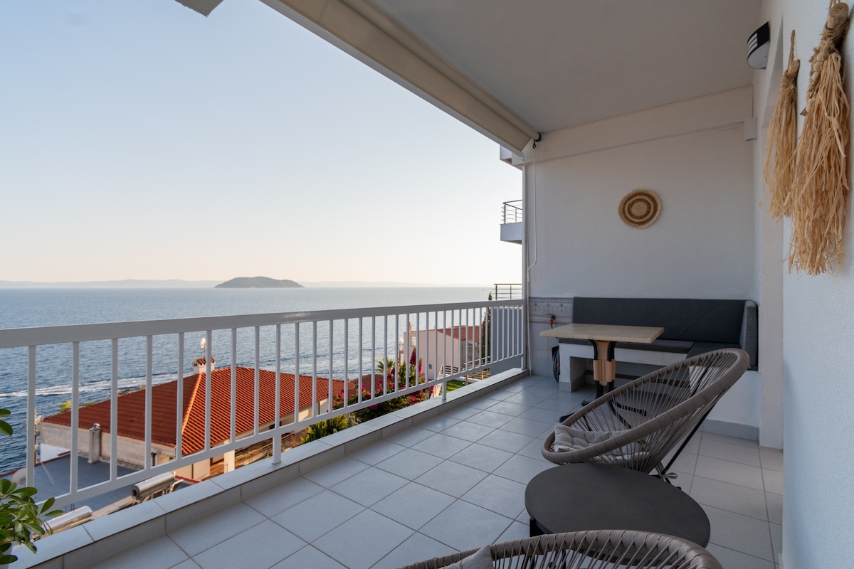 A spacious terrace offers unobstructed views of the sea and distant islands. Two lounge chairs are positioned near a seating area with a table, featuring a relaxed atmosphere. The surrounding white railing enhances the open feel, allowing natural light to flow into the space.