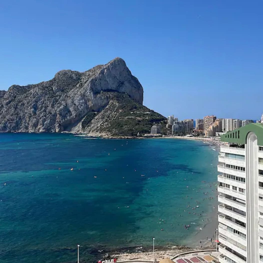 A panoramic view showcases a clear turquoise sea meeting a sandy beach, framed by a prominent rocky cliff. Modern buildings line the shore, and beachgoers can be seen enjoying the sun. The sky is bright and blue, contributing to a serene coastal atmosphere.
