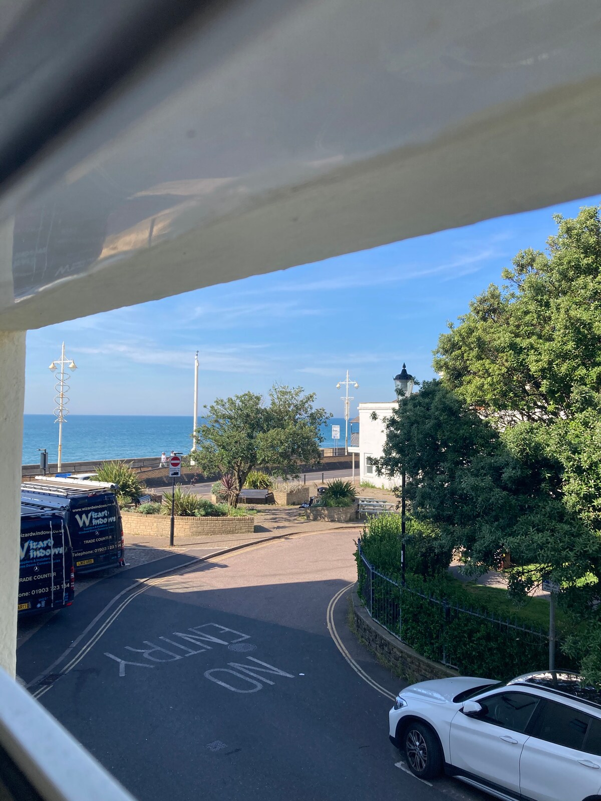 A view from a window captures the coastline of Bognor Regis. The blue ocean stretches into the distance, framed by green foliage and a charming street. Utility vehicles and street lamps are visible, blending with the town's seaside atmosphere.