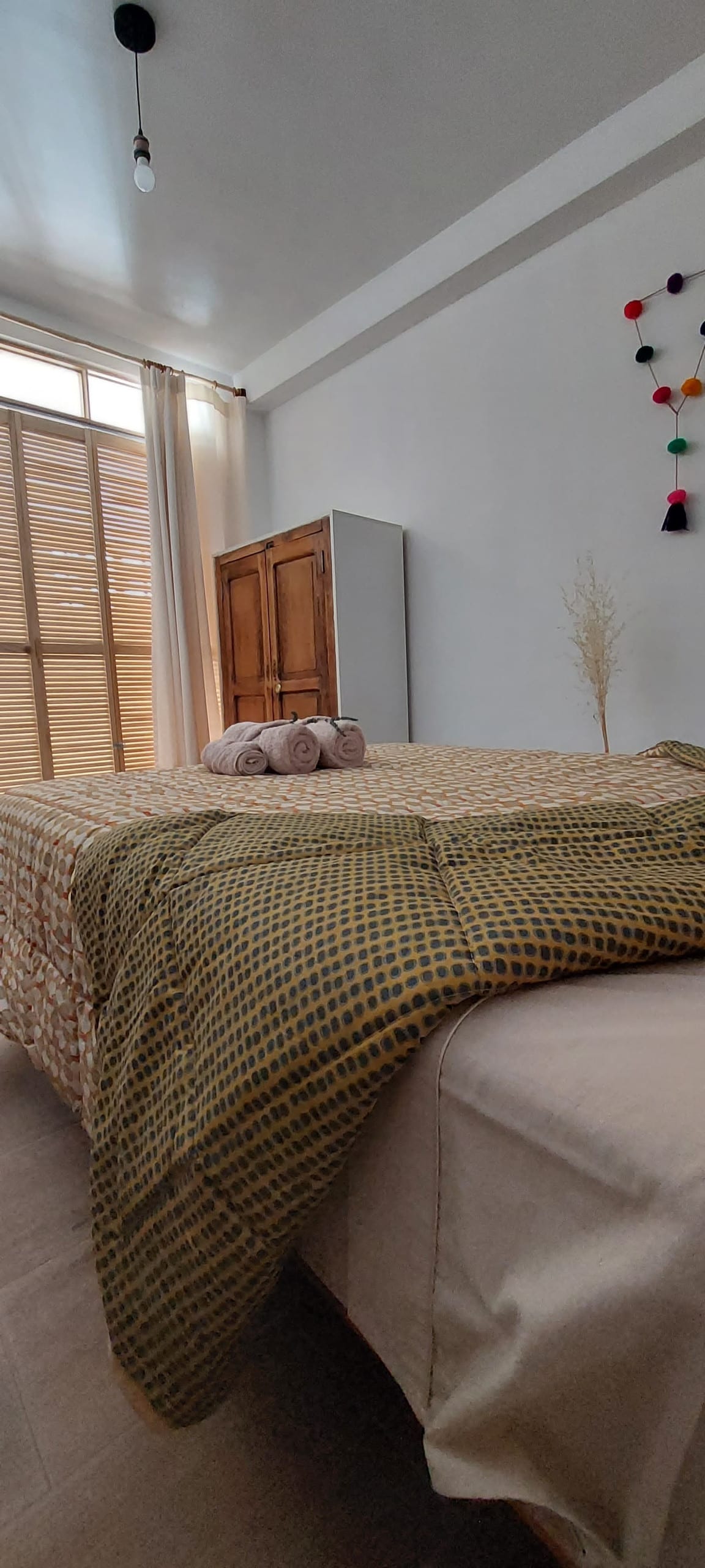 A spacious sleeping area features a textured blanket draped over the bed. Soft towels are neatly placed on top, while natural light filters in through large windows adorned with light curtains. A wooden cabinet is positioned against the wall, accompanied by decorative elements in the background.