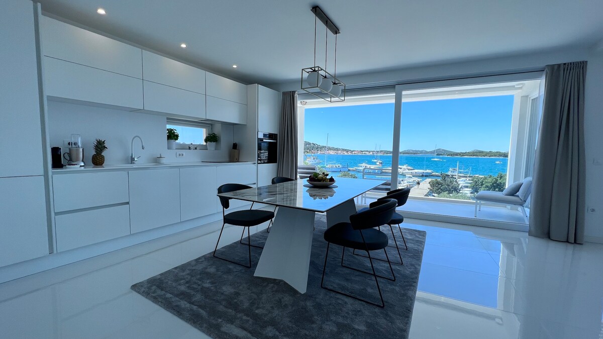 A modern kitchen and dining area is positioned beside expansive glass doors, offering a clear view of the marina and sea. A sleek dining table is surrounded by black chairs, while the kitchen features minimalist white cabinetry and a large window that allows natural light to fill the space.