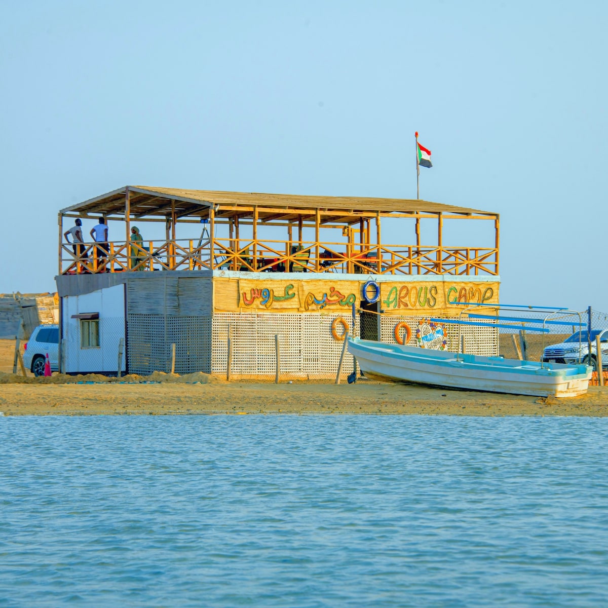 A wooden structure named Arous Camp is visible near the shoreline, featuring a welcoming sign and a flag. People can be seen on the open deck, enjoying the coastal view. A boat is anchored nearby, while the sandy beach extends along the calm water.