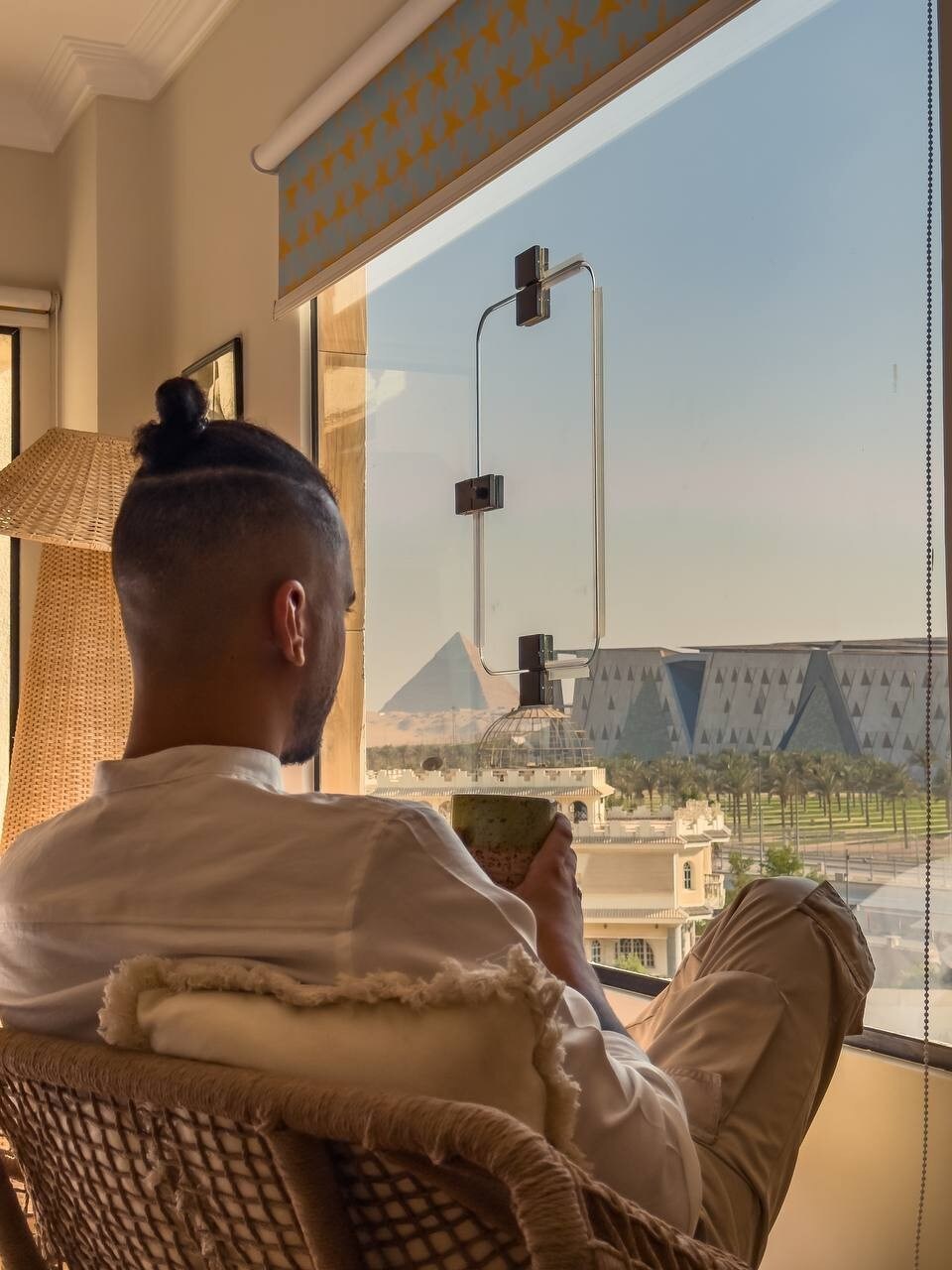 A person is seated in a woven chair, sipping from a cup, with a view of the Great Pyramids and the Grand Egyptian Museum in the background. Bright light enters through the window, accentuating the warm interior decor.
