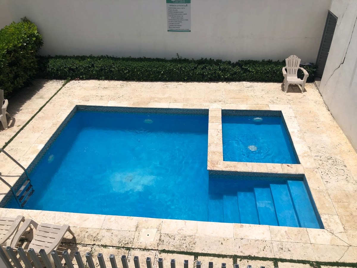 A swimming pool is set in a stone-tiled area, featuring a built-in spa section and stairs leading into the water. Lounge chairs are positioned nearby, surrounded by green foliage and a clean, white wall.
