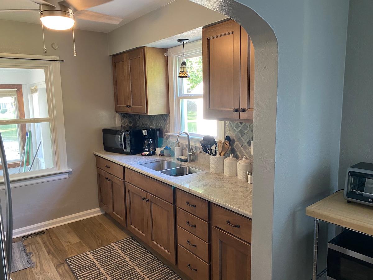 A modern kitchen features wooden cabinets and a stone countertop with a deep sink. Small kitchen appliances, utensils, and dishware are neatly arranged. Natural light enters through a nearby window, which provides a view of the outdoor area.