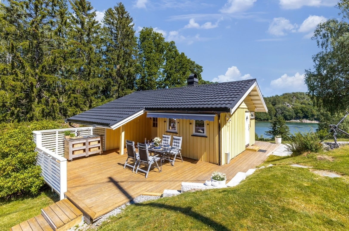 A summer cottage is positioned on a spacious deck surrounded by greenery, featuring a seating area with chairs and a table. The sloped roof and colorful exterior complement the serene coastal setting. Scenic views of the waterfront are visible in the background.