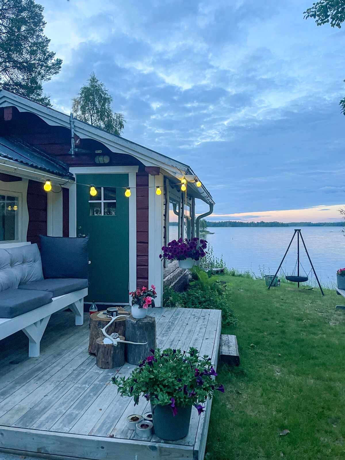 The exterior of a charming wooden cottage is shown at twilight, with soft lighting from hanging bulbs. A small deck hosts a seating area with gray cushions, while flower pots add color. The serene lake is visible in the background, reflecting the evening sky.