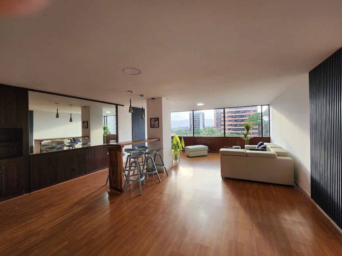 A spacious living area is highlighted by large windows that provide city views. A comfortable white sofa and a small lounge area offer relaxation options. A bar table with high chairs is positioned near the kitchen, enhancing the communal space.