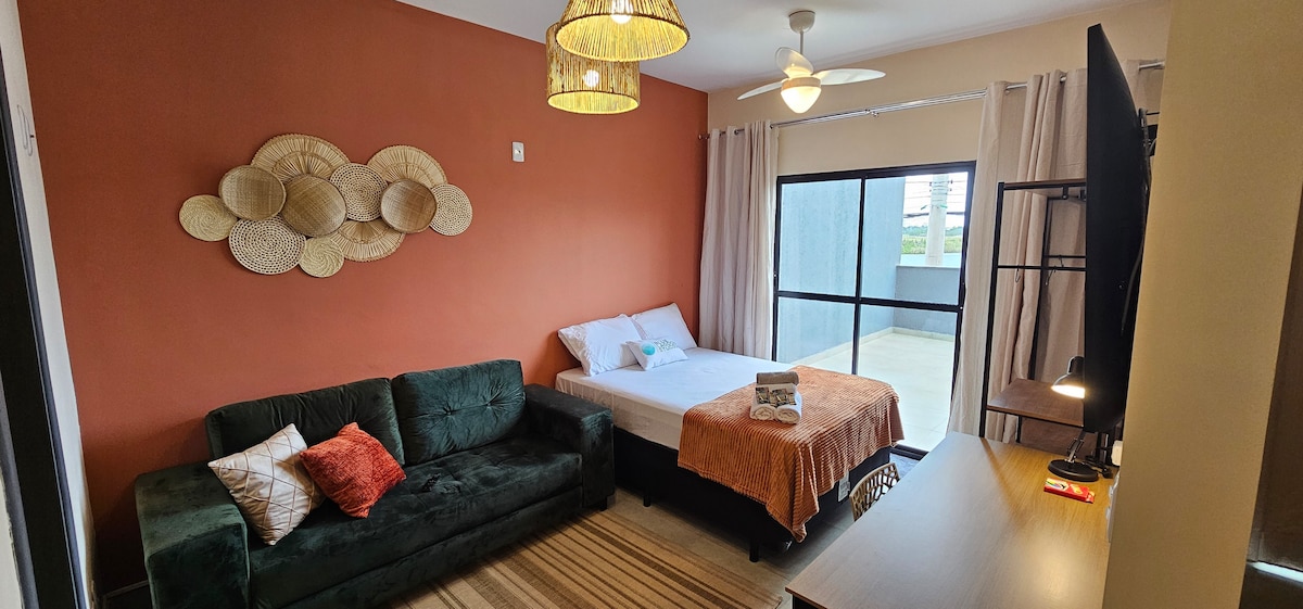 A well-lit studio is presented with a spacious bed covered in neutral linens and an inviting sofa. Textured wall decor adds a touch of warmth. A large window allows natural light to brighten the room, complemented by practical furnishings and a smart TV.
