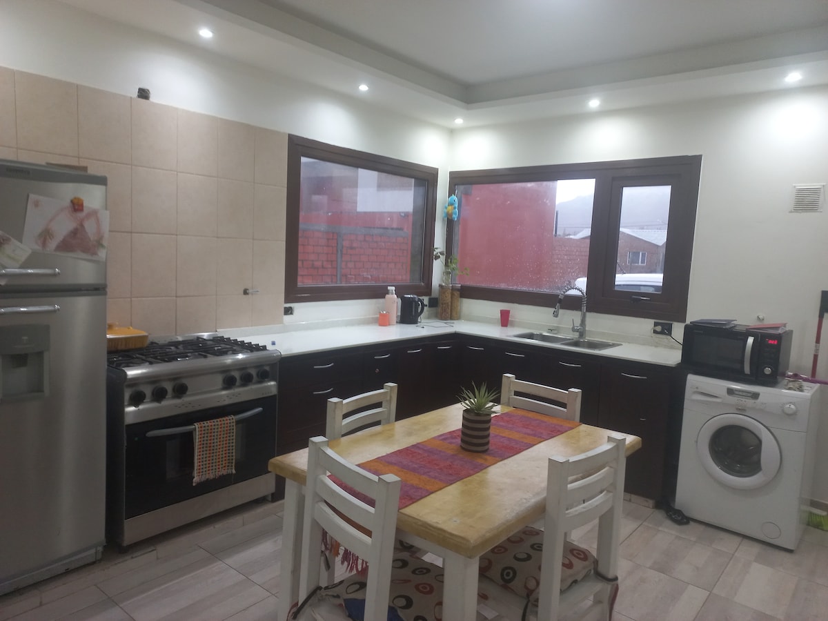 The kitchen features a modern design with light-colored tile walls and ample natural light from large windows. A wooden dining table is paired with four white chairs, while stainless steel appliances, including a gas stove and washing machine, are integrated into the space.