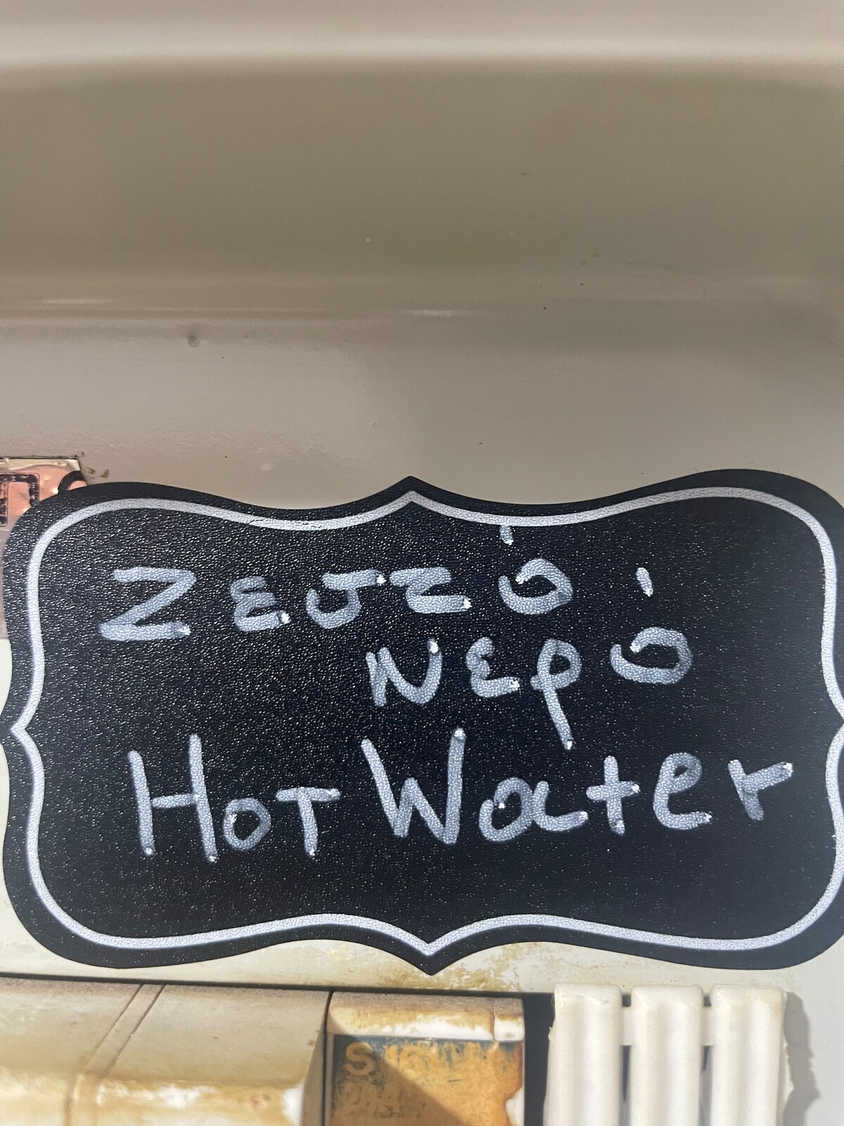 A black chalkboard sign displaying the words 'Hot Water' in white chalk. The edges of the sign are decorated with a curved border. The background features light-colored surfaces, suggesting it is mounted in a kitchen area.