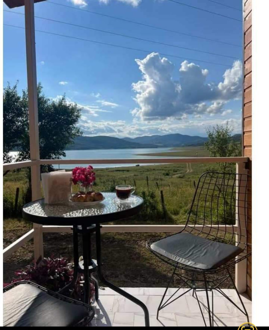 A small outdoor seating area features a round table with two chairs and is adorned with a pink flower centerpiece. Scenic views of the lake and distant mountains are visible in the background, complemented by a clear blue sky with clouds.