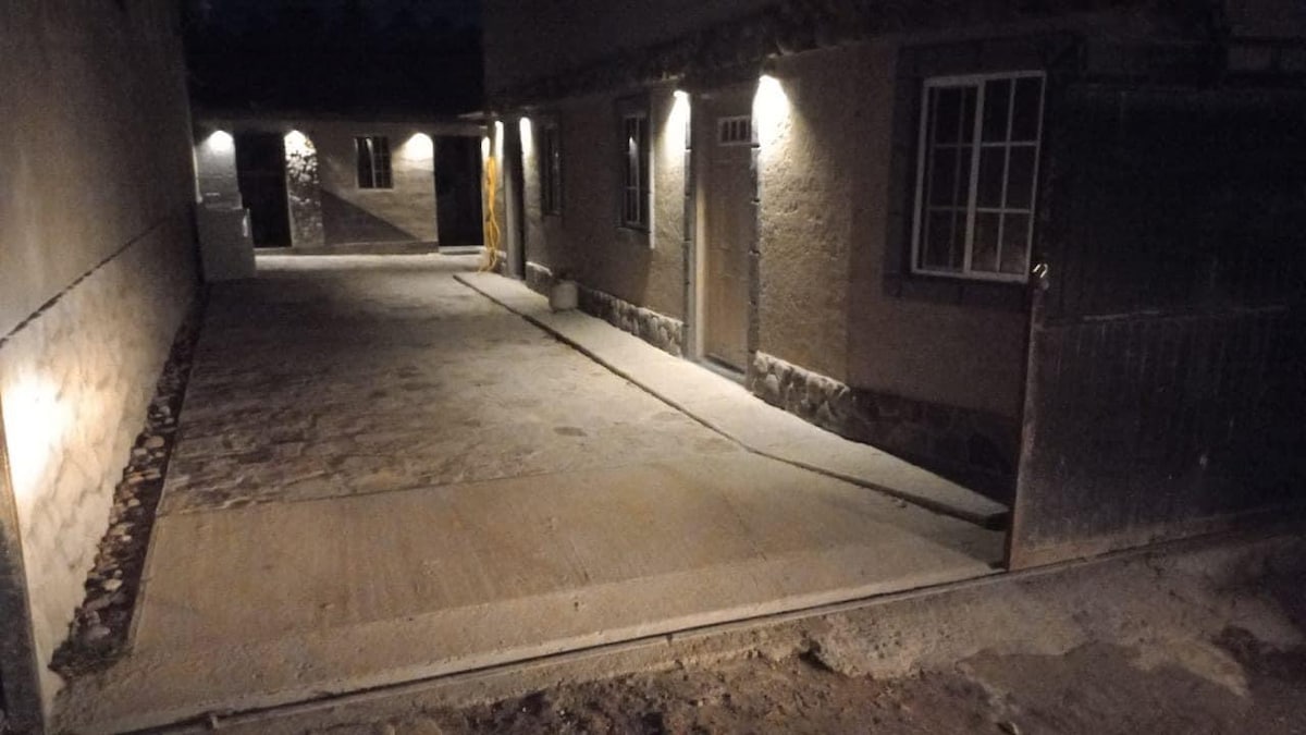 A well-lit entrance area is visible, featuring a smooth concrete path leading to a doorway. Stone walls and warm lighting create a secure and welcoming ambiance. Additional windows are seen along the structure, enhancing the spaciousness of the entryway.
