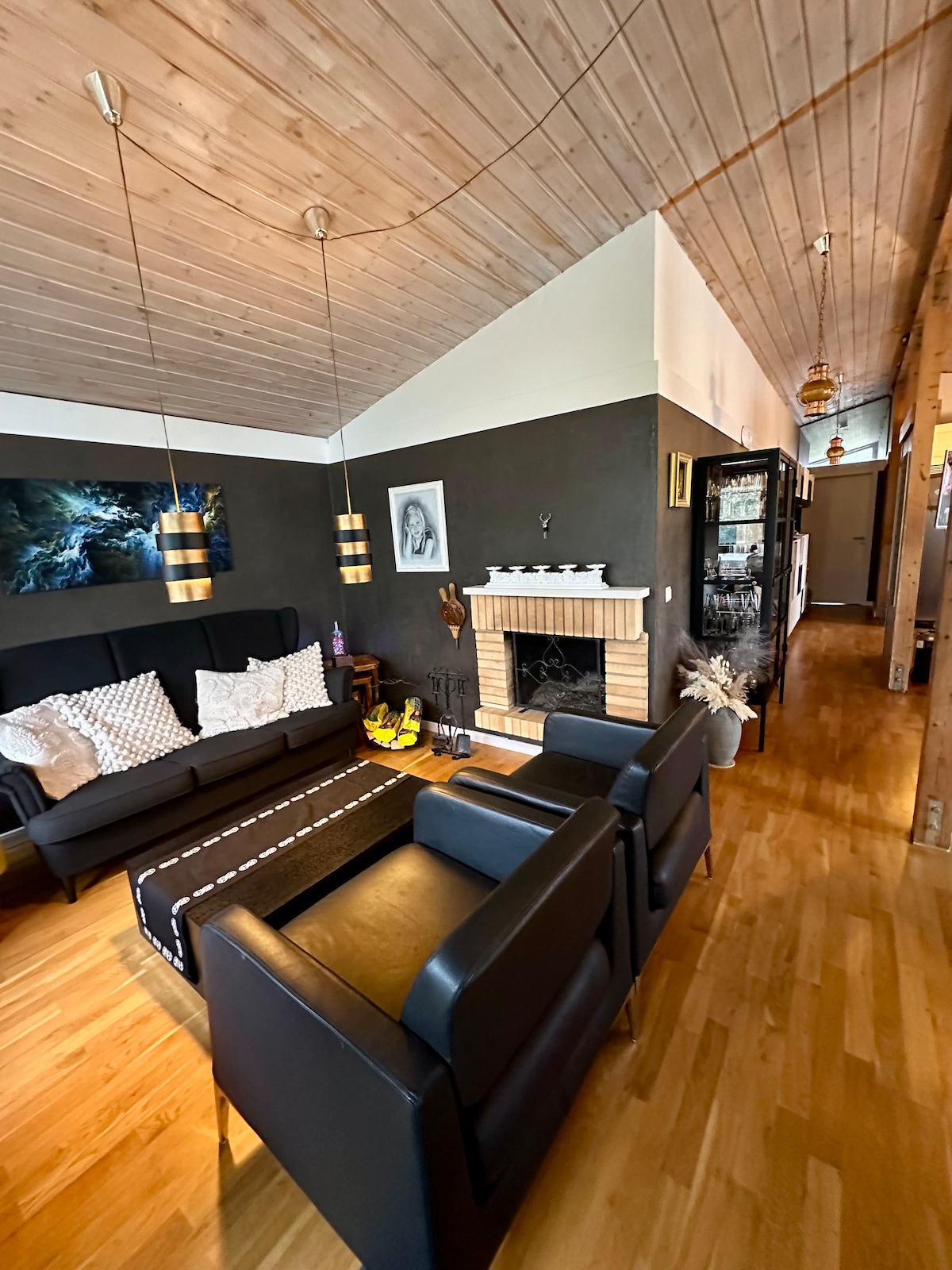 A spacious living area features a black sectional sofa and armchairs, arranged around a central coffee table. A fireplace with a decorative mantel adds warmth to the room. Warm wood accents and high ceilings contribute to an inviting atmosphere, while artwork adorns the walls.