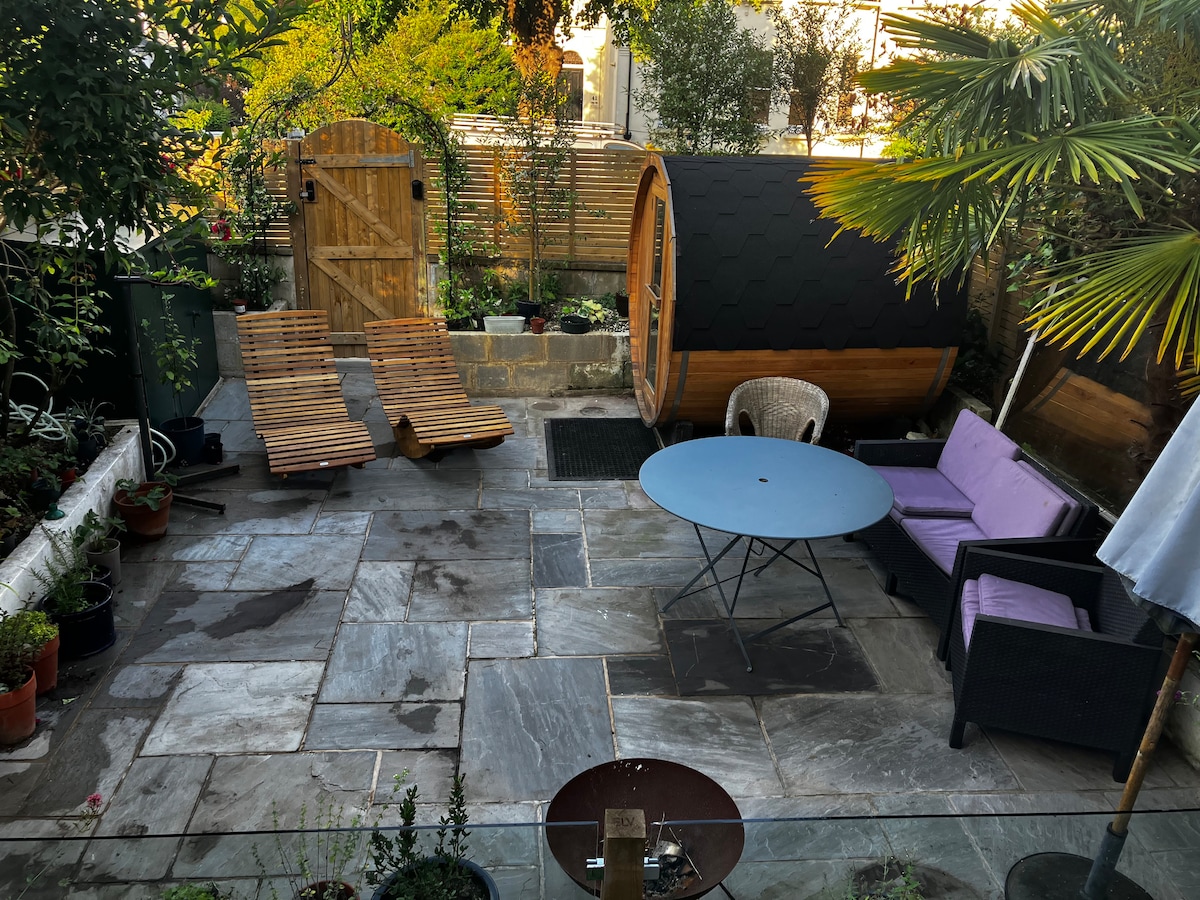 A private garden is featured, with natural stone flooring and various potted plants. Lounge chairs in wood are positioned alongside a round table, while outdoor seating in black wicker and purple cushions is visible. An electric sauna is located toward the back, surrounded by greenery.