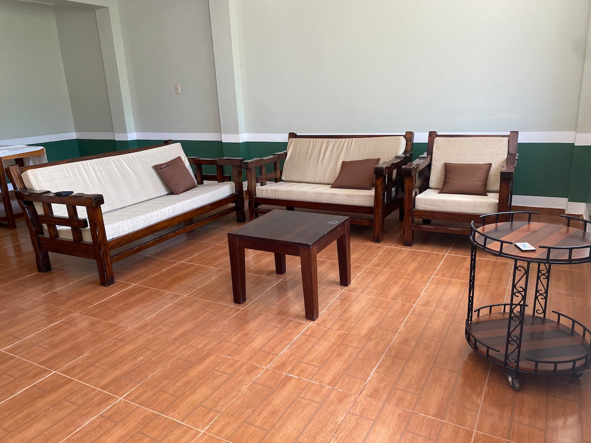A spacious seating area is presented with a combination of wooden sofas, featuring light-colored cushions. A small wooden coffee table occupies the center, complemented by a circular side table with a dark finish. The tile floor adds a warm tone to the space.