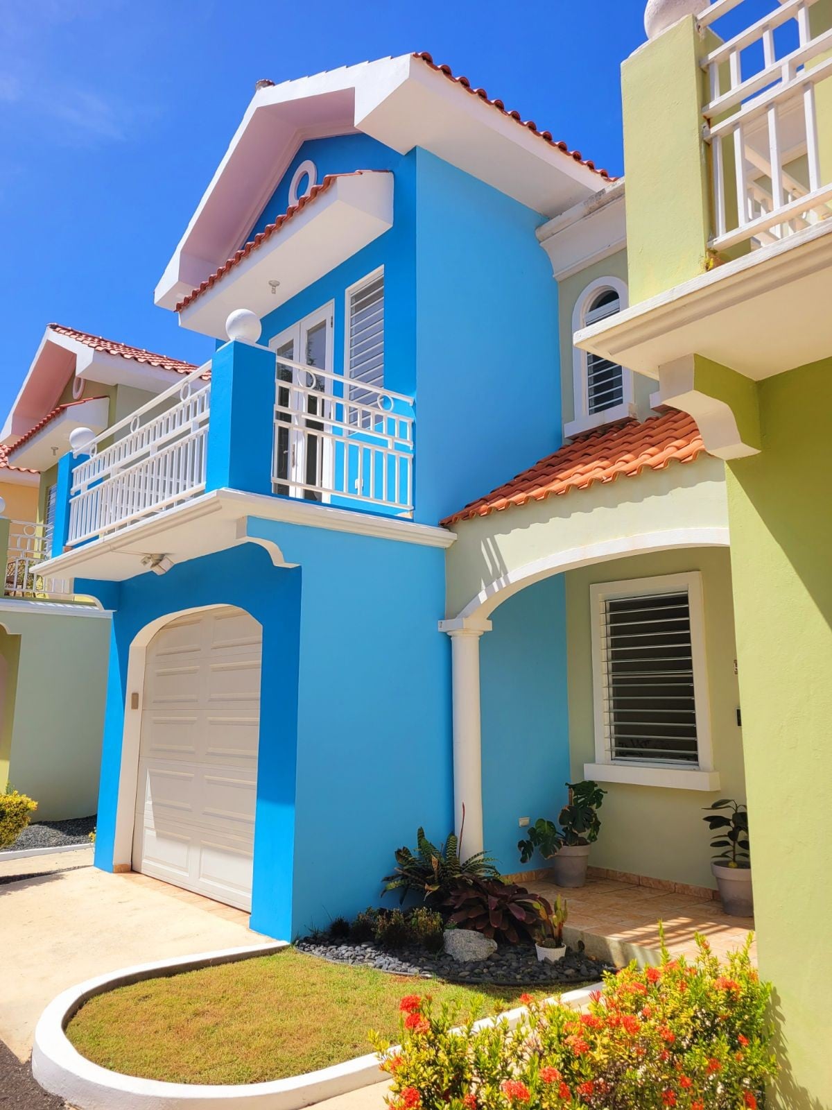 A vibrant two-story condo is depicted, featuring a light blue exterior with a red-tiled roof. A white garage door is visible, along with a small garden area that includes various plants and flowers. The bright sky enhances the inviting appearance of the home.