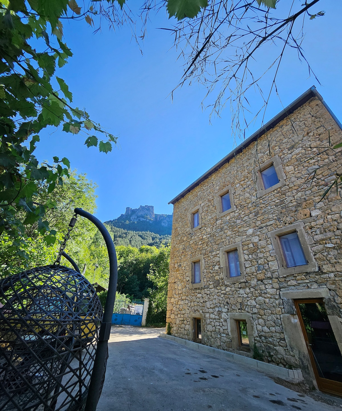 The exterior of a stone building is presented against a clear blue sky. Surrounded by greenery, a sculptural hanging chair is visible in the foreground. The building showcases large windows and a rugged stone facade, blending harmoniously with the natural landscape and the distant mountainous backdrop.
