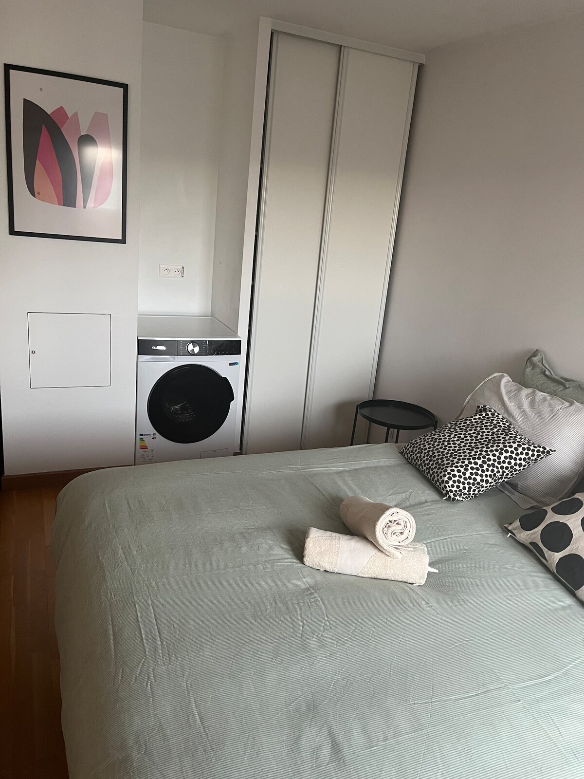 A comfortable sleeping area features a spacious bed dressed in soft, light-colored linens. A black round side table is positioned nearby, and a washing machine is tucked into a designated space. Decorative pillows add a subtle touch of style, complemented by framed wall art.