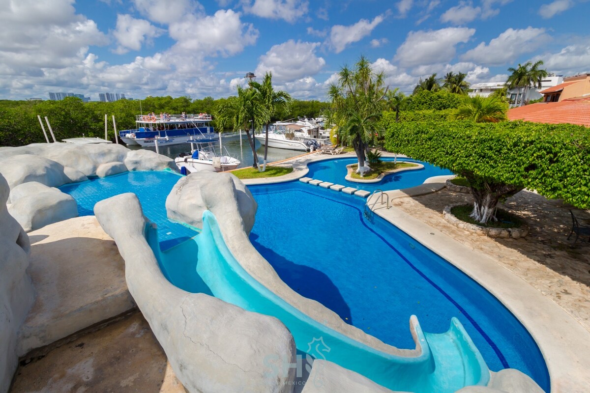 A unique outdoor pool area is surrounded by lush greenery, featuring water slides integrated into a rock-like structure. The pool's vibrant blue water reflects the sky, while lounge chairs are positioned nearby, providing a relaxing space for guests.