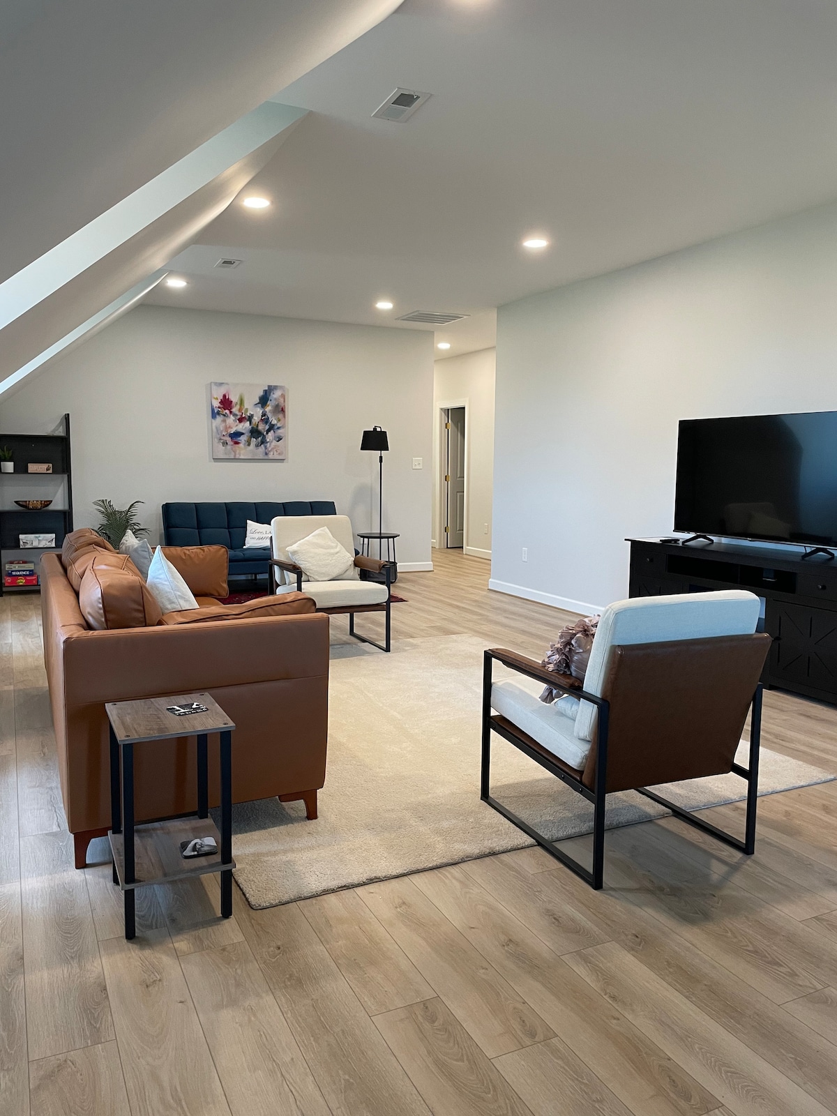 The living room features a mix of seating options, including a brown leather sofa, two armchairs, and a side table. A television is positioned on a dark console across from the seating. Soft lighting brightens the space, complemented by wooden flooring and light-colored walls.