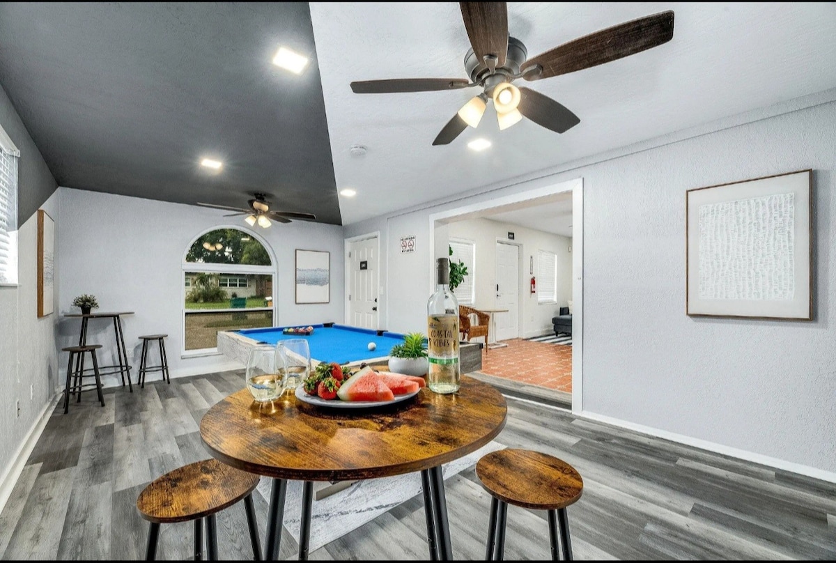 A room features a dining area with a round table and three stools. A beverage and sliced fruit are placed on the table. A pool table is visible in the background, with an arched window allowing natural light to enter the space.