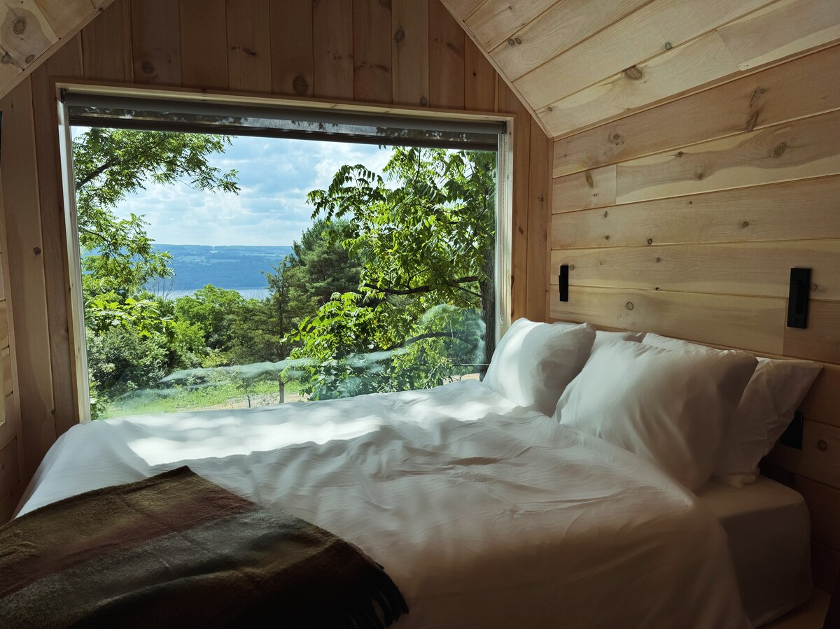 A cozy sleeping area is framed by a large window, offering views of lush greenery and Seneca Lake in the distance. The spacious bed is adorned with crisp white linens, complemented by a soft throw blanket at the foot.