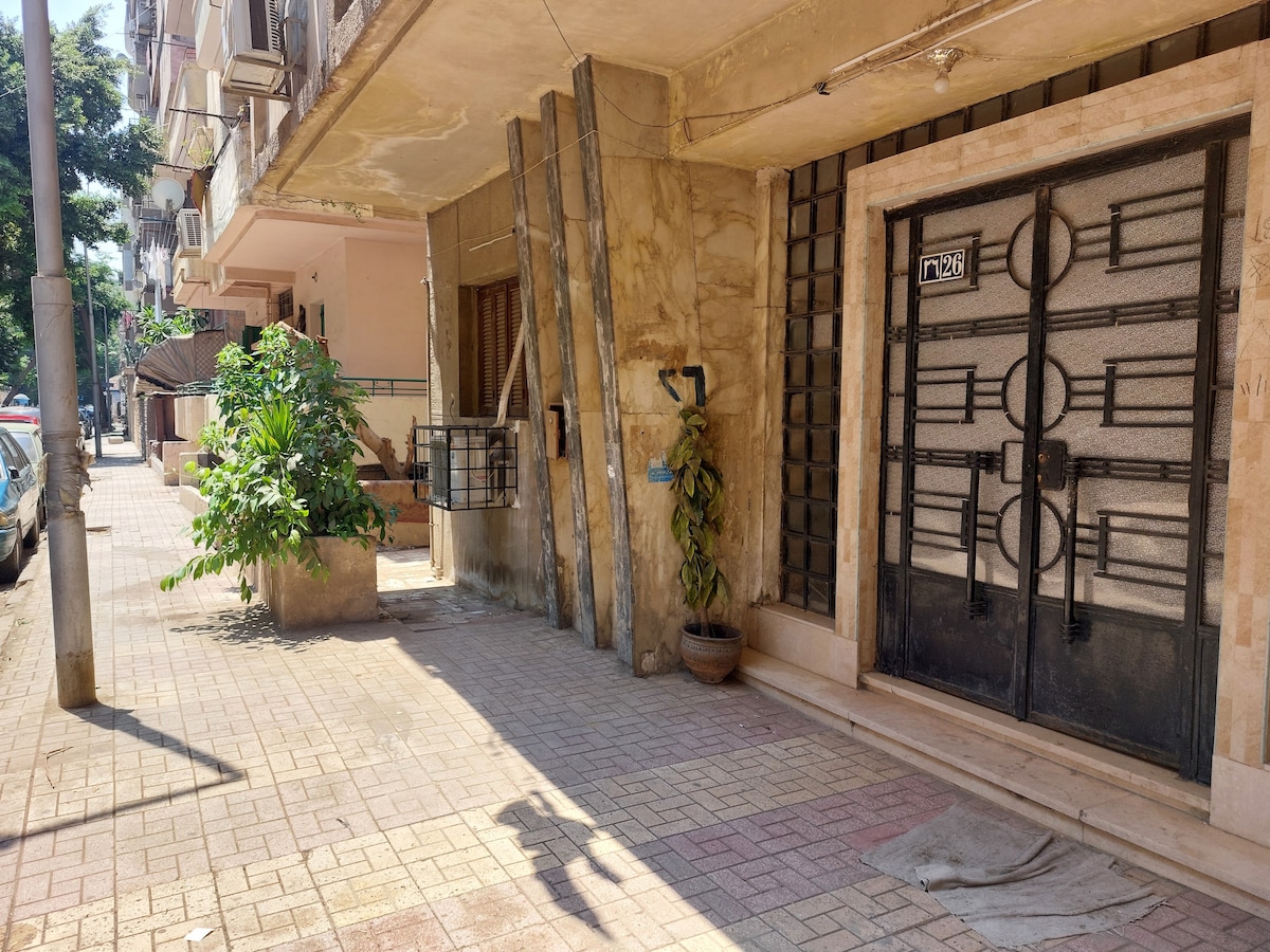 The image shows the entrance area of an apartment building on a calm street. A large black metal gate leads to the entrance, flanked by a green plant in a pot. Sidewalks and other buildings are visible in the background.