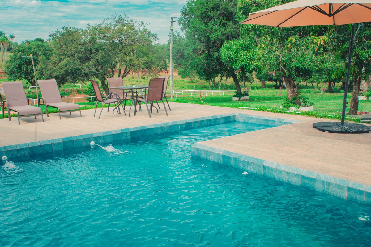 A refreshing swimming pool is framed by lush greenery, featuring several sun loungers and a dining table underneath a large umbrella. The pool area offers a peaceful retreat, surrounded by well-maintained grass and trees, inviting relaxation and outdoor activities.