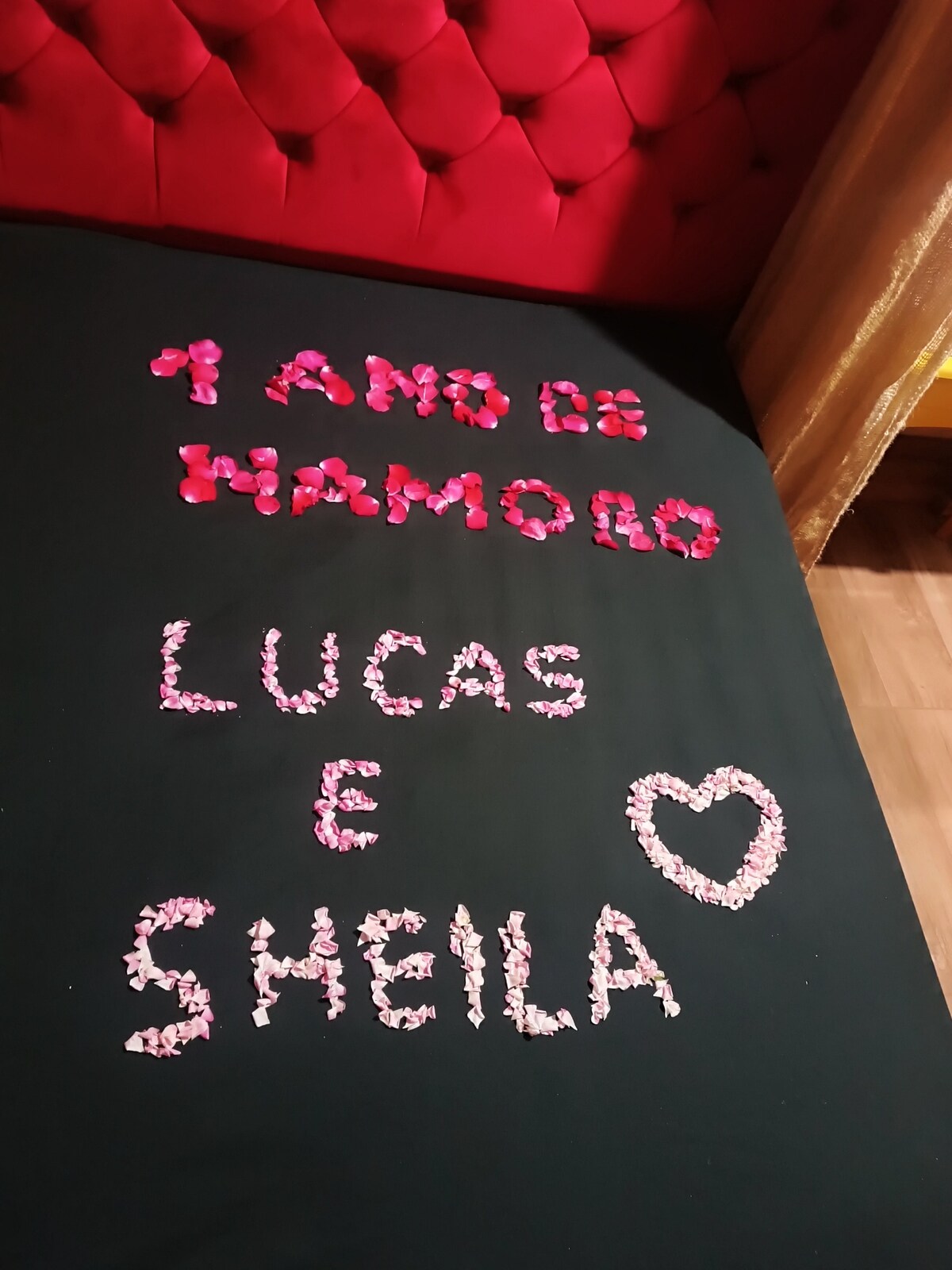 A bed with a dark cover features petals arranged in a message reading '1 ANO DE NAMORO' and the names 'LUCAS E SHEILA' in varying shades of pink. A heart shape is also created with petals, adding a decorative touch.