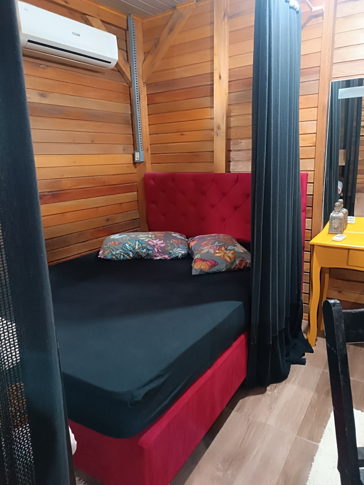 A cozy sleeping area is featured, with a fitted black bedspread covering a spacious red upholstered bed. Two decorative pillows with floral patterns add color. Dark curtains frame the entrance, while a wooden wall provides a warm backdrop. An air-conditioning unit is mounted above.