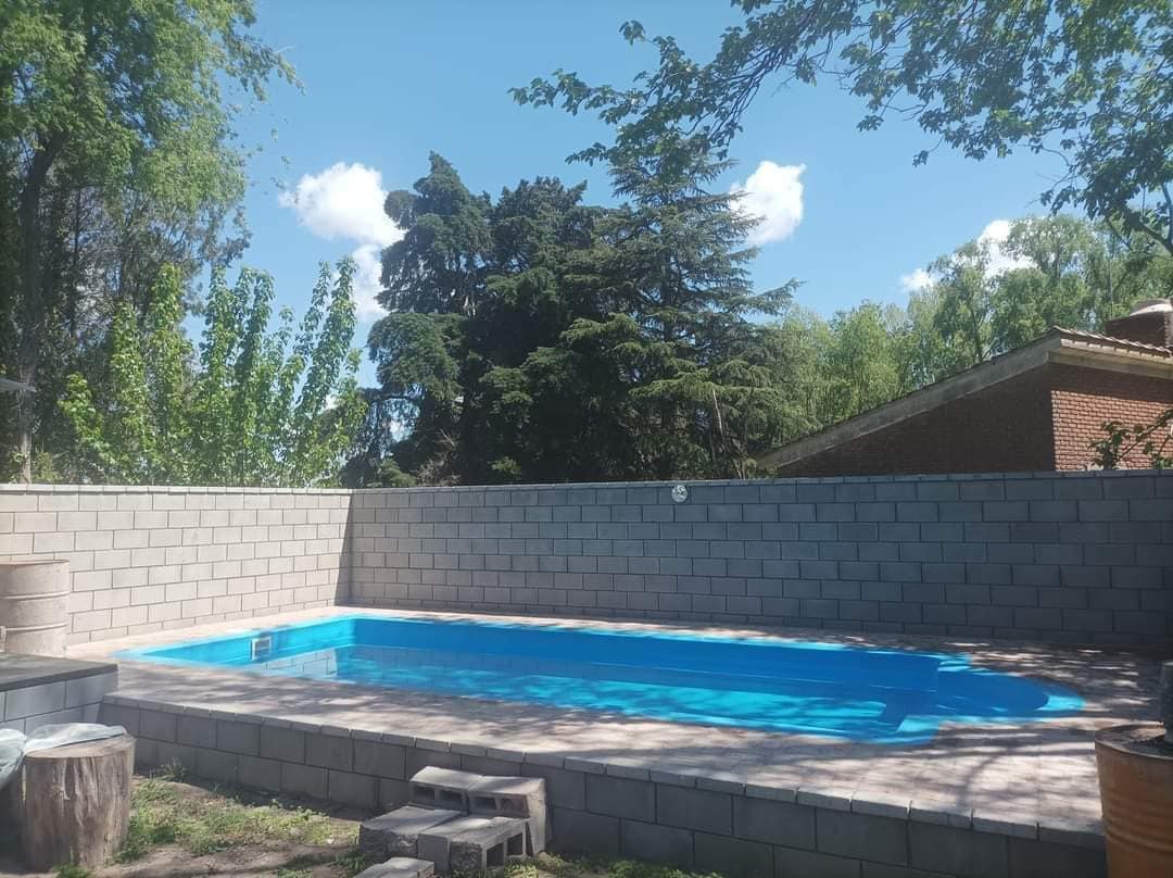 A rectangular swimming pool is set within a stone patio, surrounded by greenery and tall trees. A light blue hue of the water reflects the clear sky and scattered clouds, creating a serene outdoor space for relaxation.
