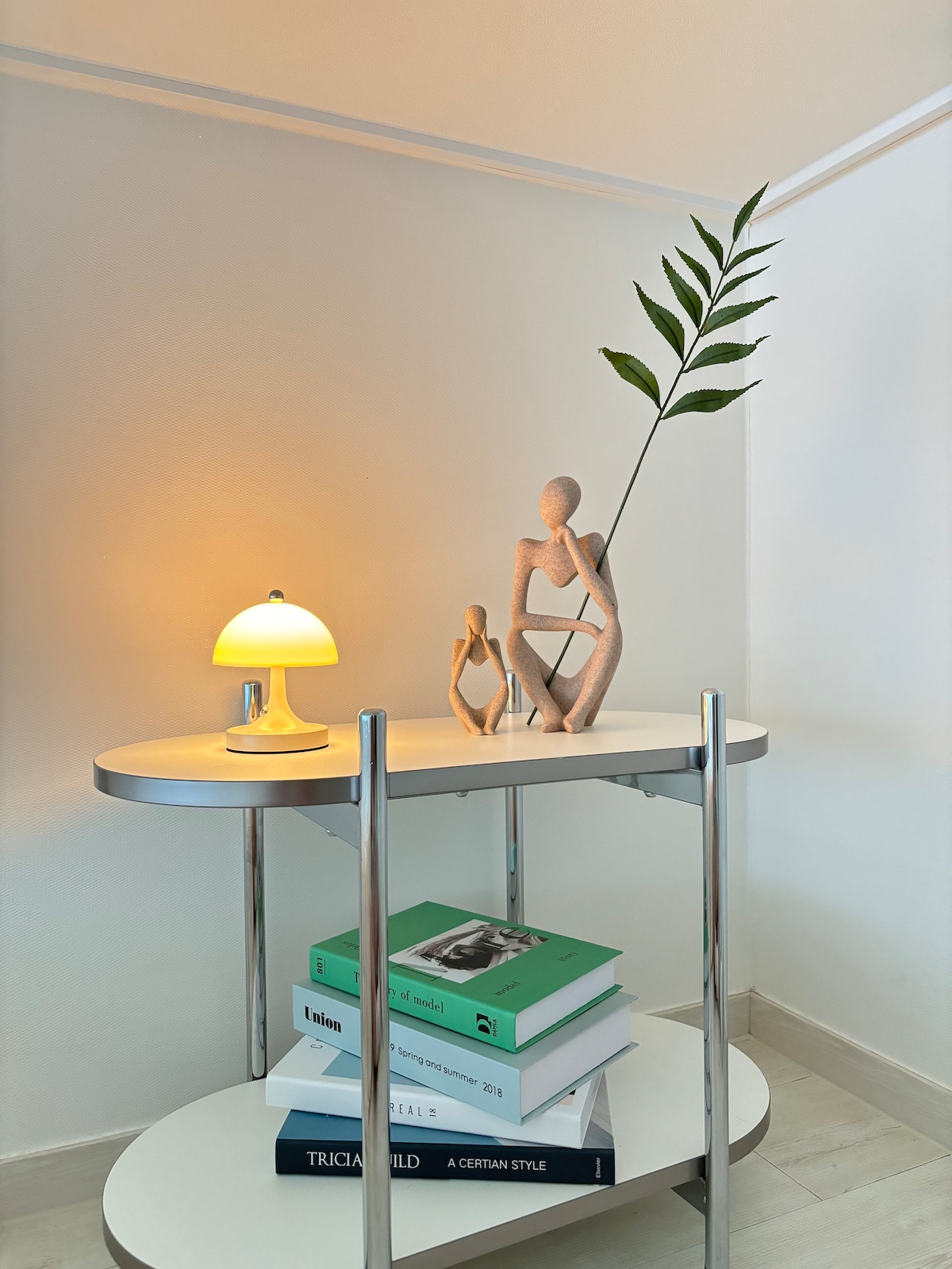 A sleek white side table is positioned against a light wall. A modern lamp emits a soft glow, illuminating two decorative figurines and a green leaf in a minimalist vase. Stacked books in various colors are neatly arranged beneath the tabletop.