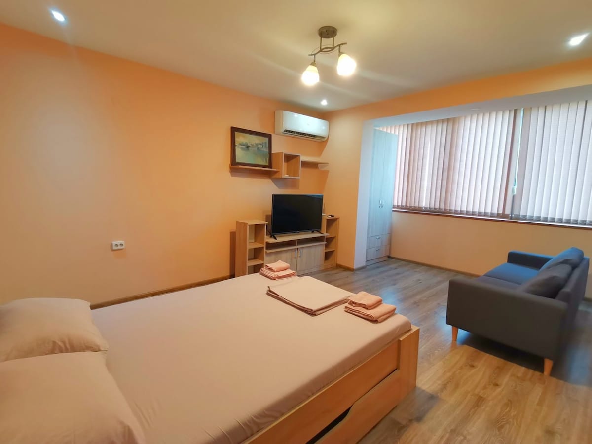 A spacious bedroom is presented with light orange walls and wooden flooring. A large bed is positioned in the foreground, while a television is mounted on a shelf across from a comfortable seating area with a gray sofa. Natural light filters through blinds on a large window.