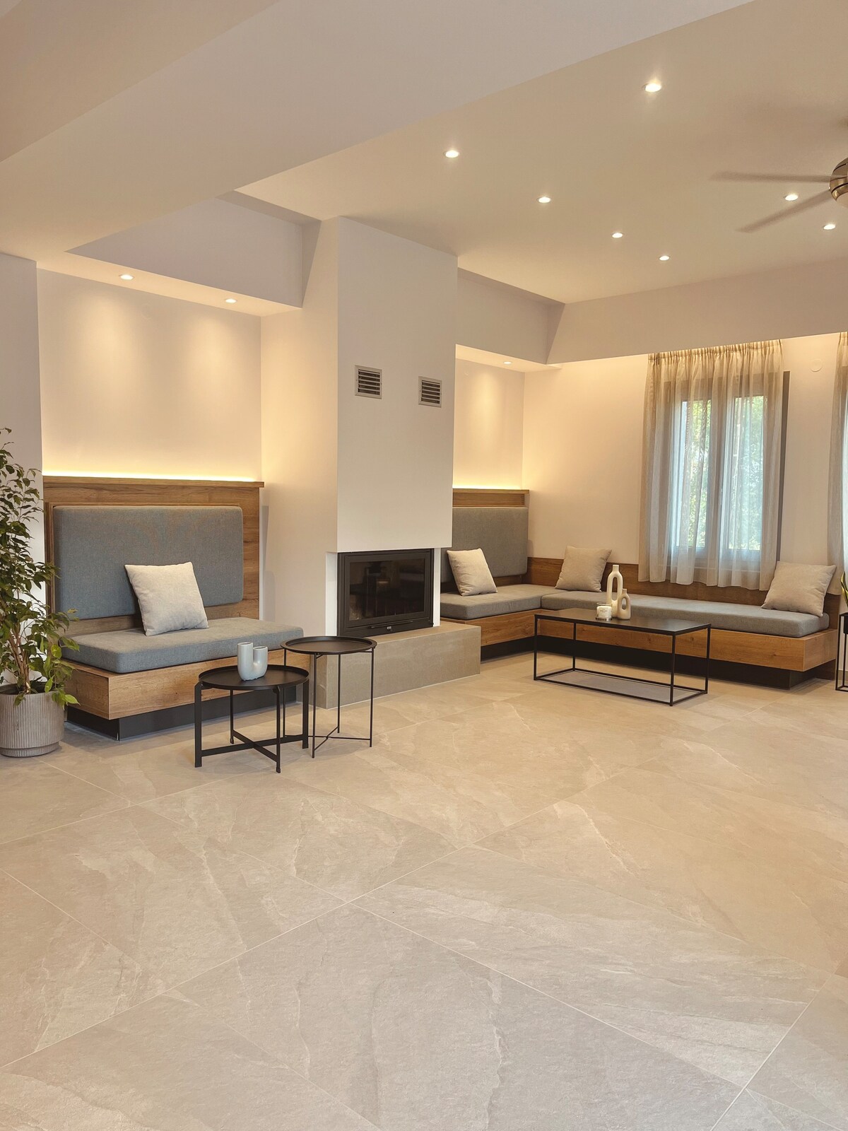 The spacious living area features soft seating against the light walls, illuminated by recessed lighting. A modern fireplace is centered in the space, with sleek tables positioned nearby. Natural light filters through the sheer curtains, enhancing the serene ambiance.