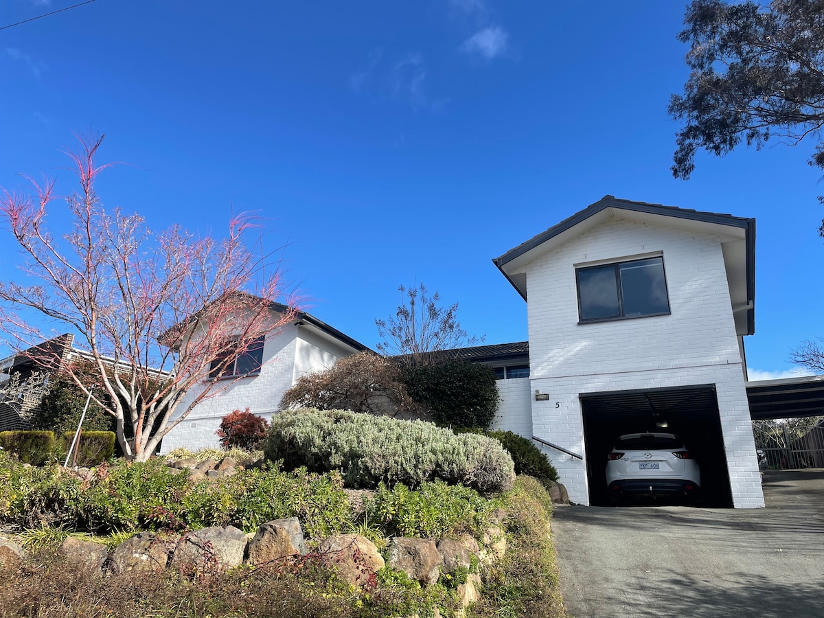 The exterior of a spacious two-story house is depicted against a clear blue sky. Well-maintained gardens feature various shrubs and a small tree in bloom. A driveway leads to a covered carport, showcasing easy accessibility.