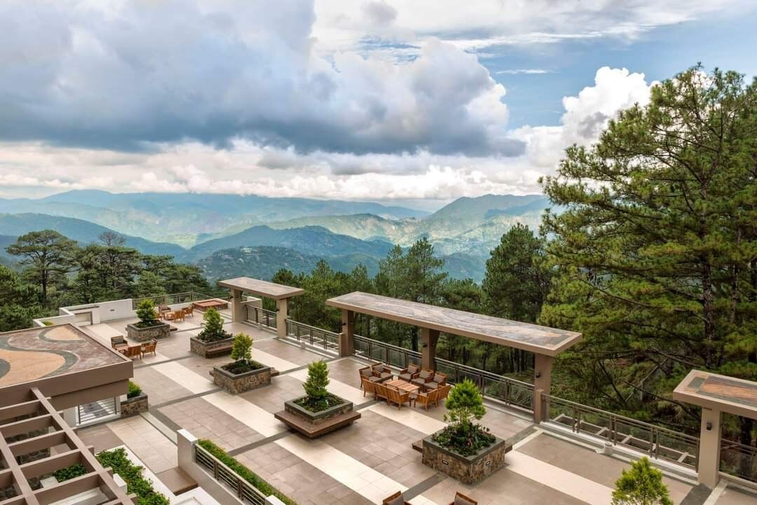 An expansive outdoor terrace offers views of rolling mountains and lush greenery. Potted plants create natural accents along the stone-paved areas, while wooden tables and chairs are arranged for gatherings. The partially cloudy sky adds depth to the serene landscape.
