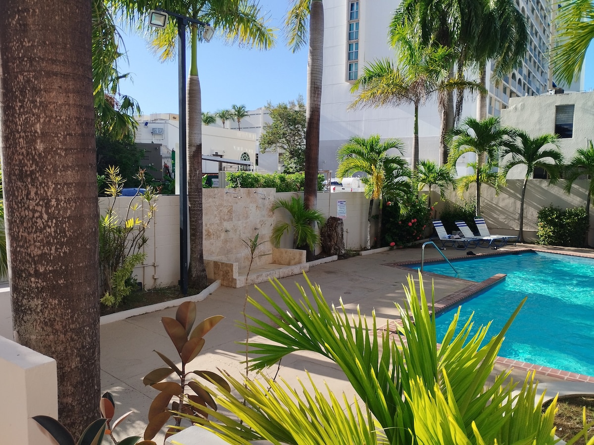 A serene pool area is surrounded by lush tropical plants and palm trees. A stone wall provides a natural backdrop, while lounge chairs are positioned near the water for relaxation. The pool glistens under sunlight, inviting guests to enjoy a refreshing swim.