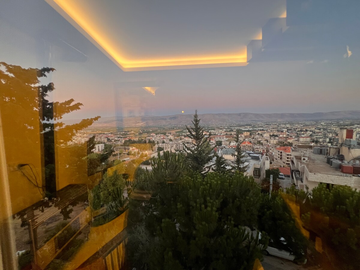 A panoramic view of the Bekaa valley is showcased through large glass windows. The landscape includes distant hills and urban areas, with greenery from surrounding trees framed in the foreground. The soft glow of ceiling lights provides a warm ambiance to the indoor space.