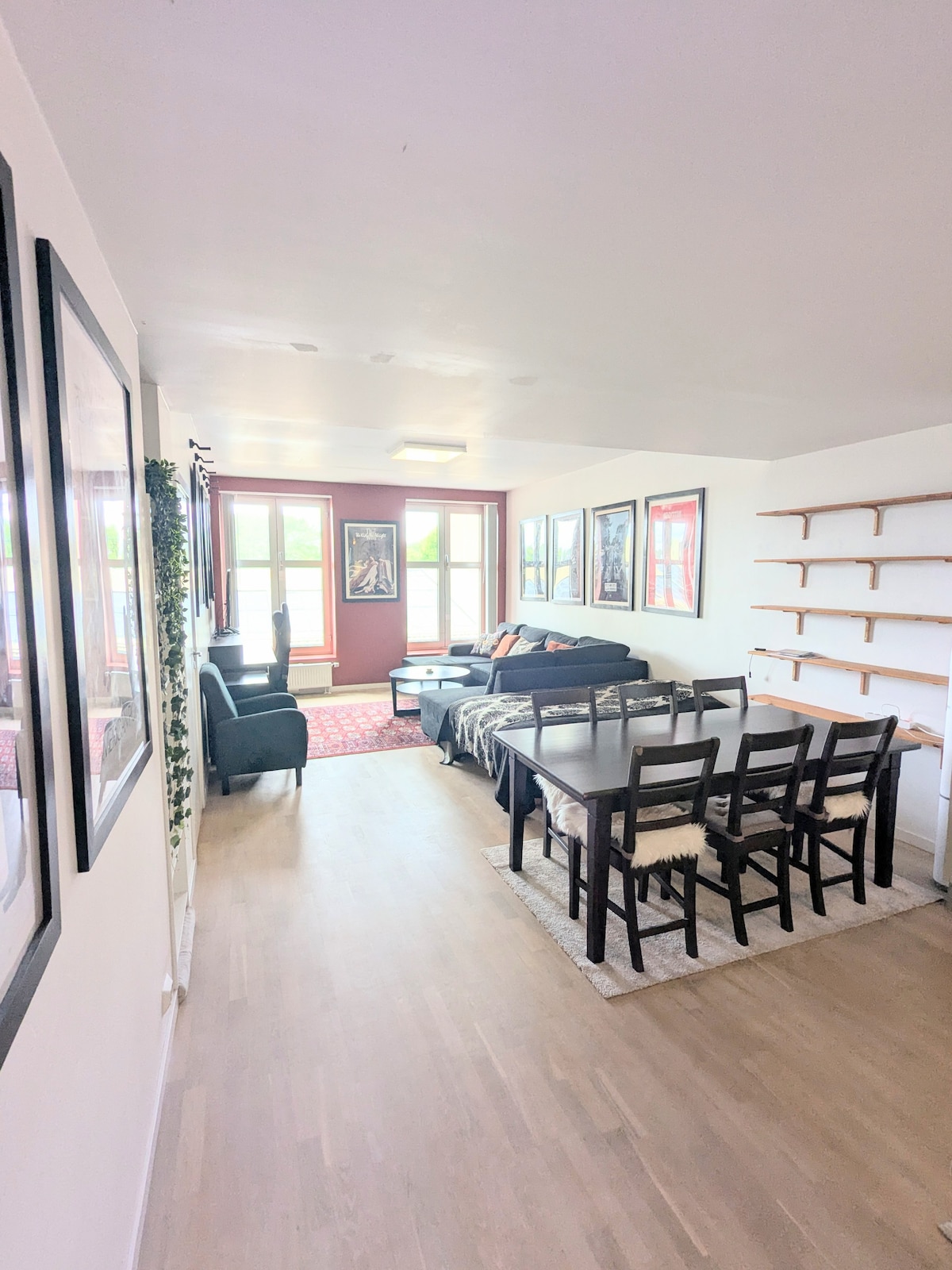 A bright living area is presented, featuring large windows allowing natural light to fill the space. A comfortable seating arrangement includes a couch and armchairs, with a dining table set for six. Wall art adds character to the clean, modern decor.