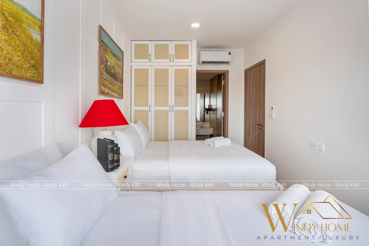 A bright and airy bedroom features two spacious beds neatly made with white linens. A red lamp adds a pop of color on the bedside table. Behind, a wooden wardrobe provides ample storage, and a doorway leads to another area of the apartment.